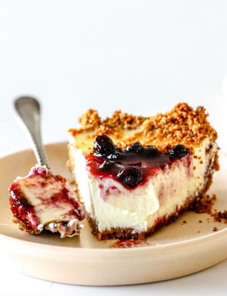 This is a side view of a slice of cream cheese pie. The slice is on a small beige plate and is topped with blueberry jam. A fork is taking off a bite of the pie and leaning on the side of the plate. The plate sits on a white counter with a white background.