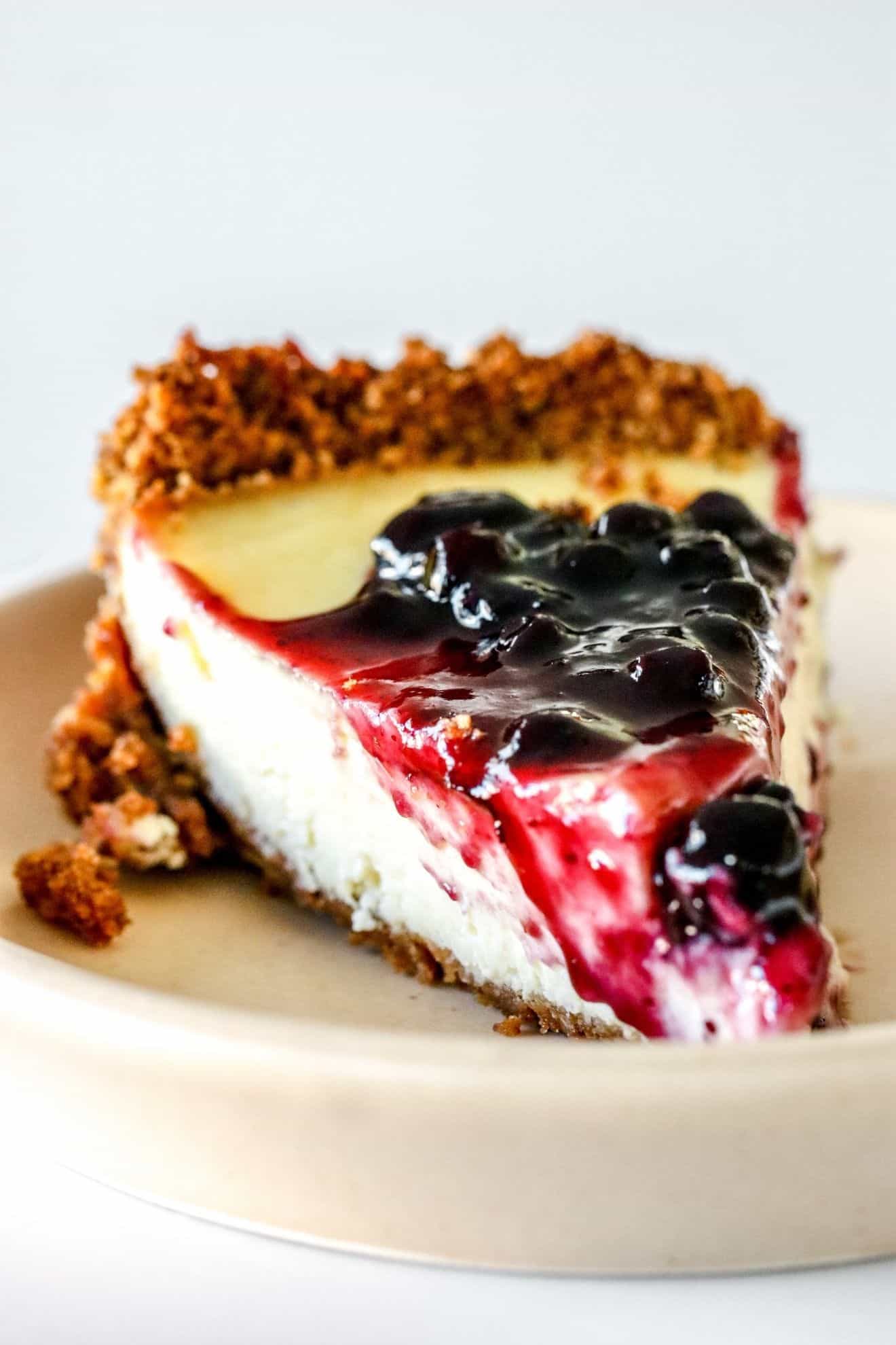 This is a side view of a slice of cream cheese pie. The slice is on a small beige plate and is topped with blueberry jam. The plate sits on a white counter with a white background.