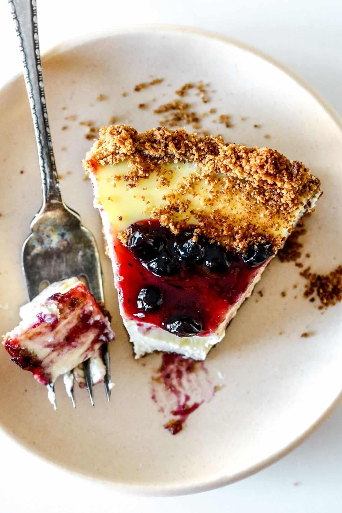 This is an overhead view of a slice of cream cheese pie. The slice is on a small beige plate and is topped with blueberry jam. A fork is taking off a bite of the pie and leaning on the side of the plate. The plate sits on a white counter with a white background.
