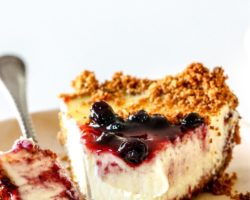 This is a side view of a slice of cream cheese pie. The slice is on a small beige plate and is topped with blueberry jam. A fork is taking off a bite of the pie and leaning on the side of the plate. The plate sits on a white counter with a white background. Text overlay reads "blueberry cream cheese pie."