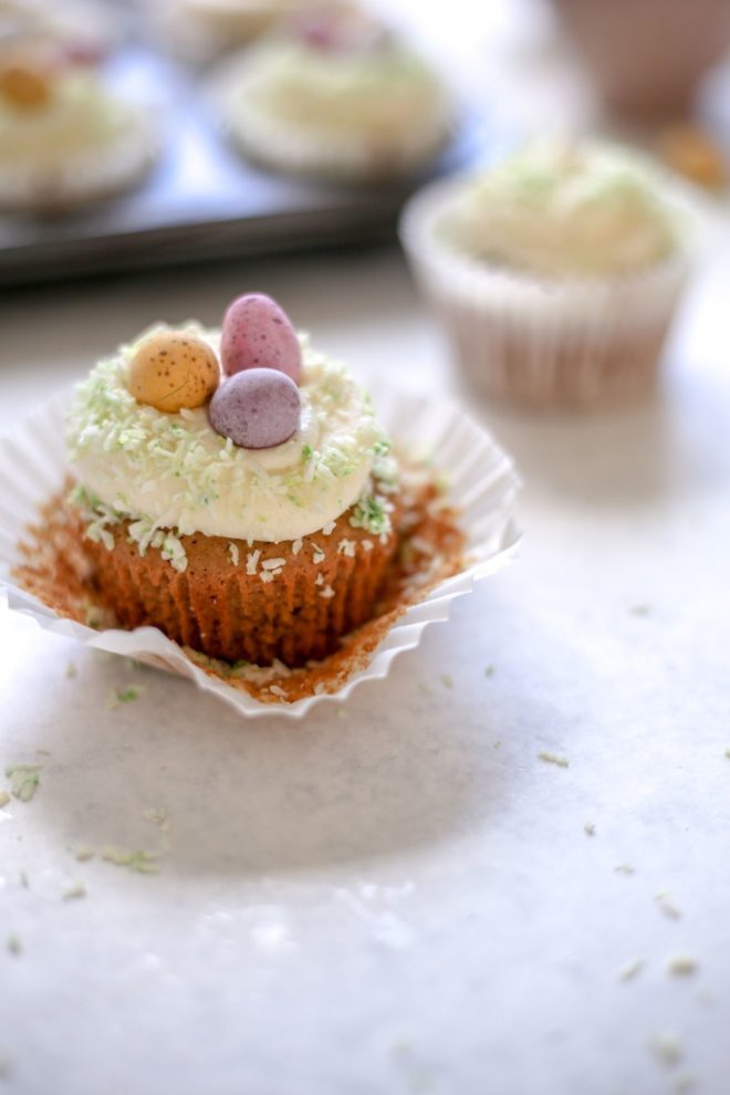 This is a side view of a vanilla cupcake with the liner peeled back. The cupcake sits on a light grey surface with more cupcakes blurred in the background. The cupcake is topped with vanilla frosting, light green coconut, and candy eggs.