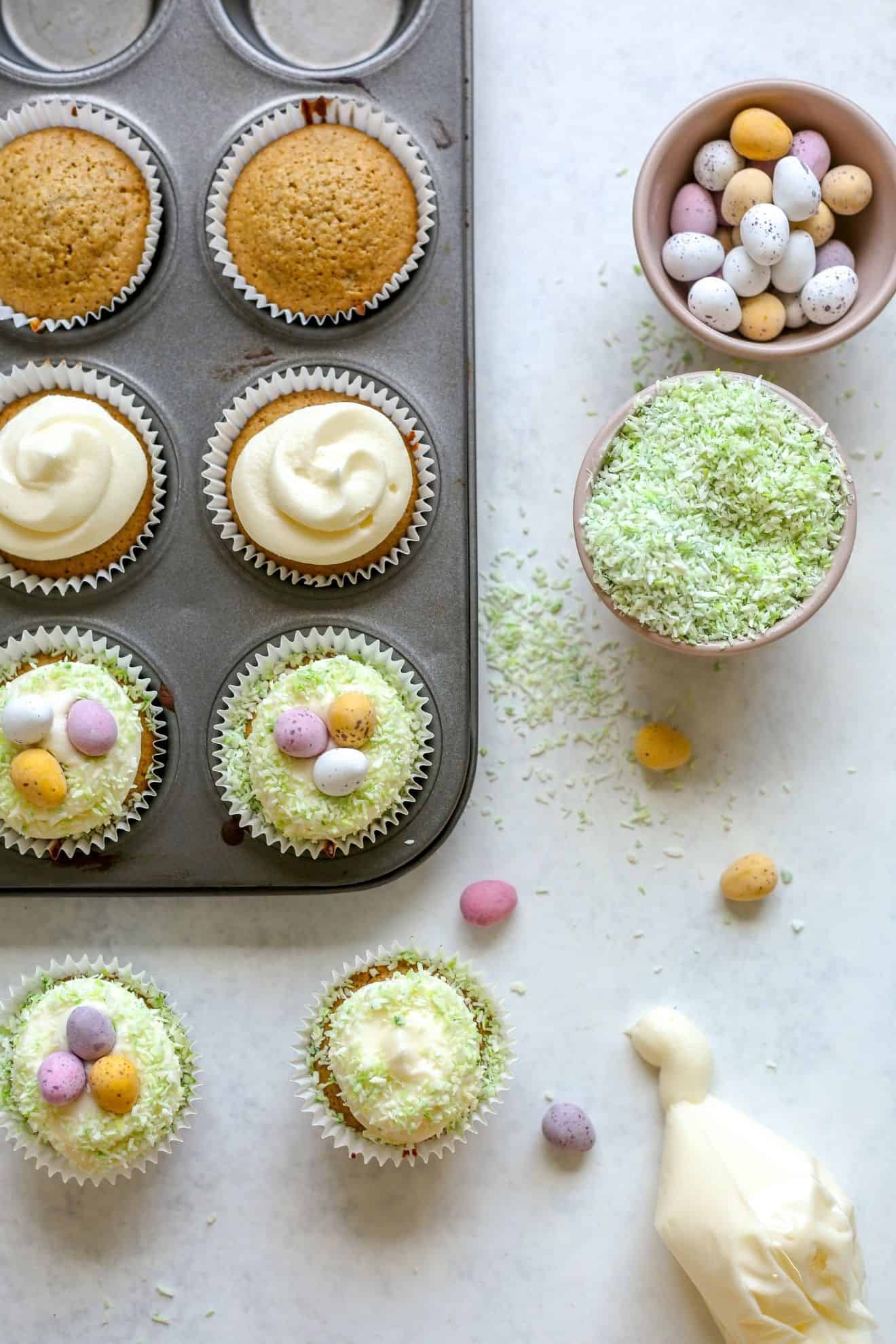 This is an overhead image of vanilla cupcakes. Some cupcakes are in the cupcake tin and some are on the white surface at the bottom of the image. The cupcakes are topped with vanilla frosting, sprinkled with green coconut and mini candy eggs. Next to the tin is a small bowl with candy eggs, and another bowl with light green colored coconut. To the bottom right of the image is a plastic piping baggie with vanilla frosting.