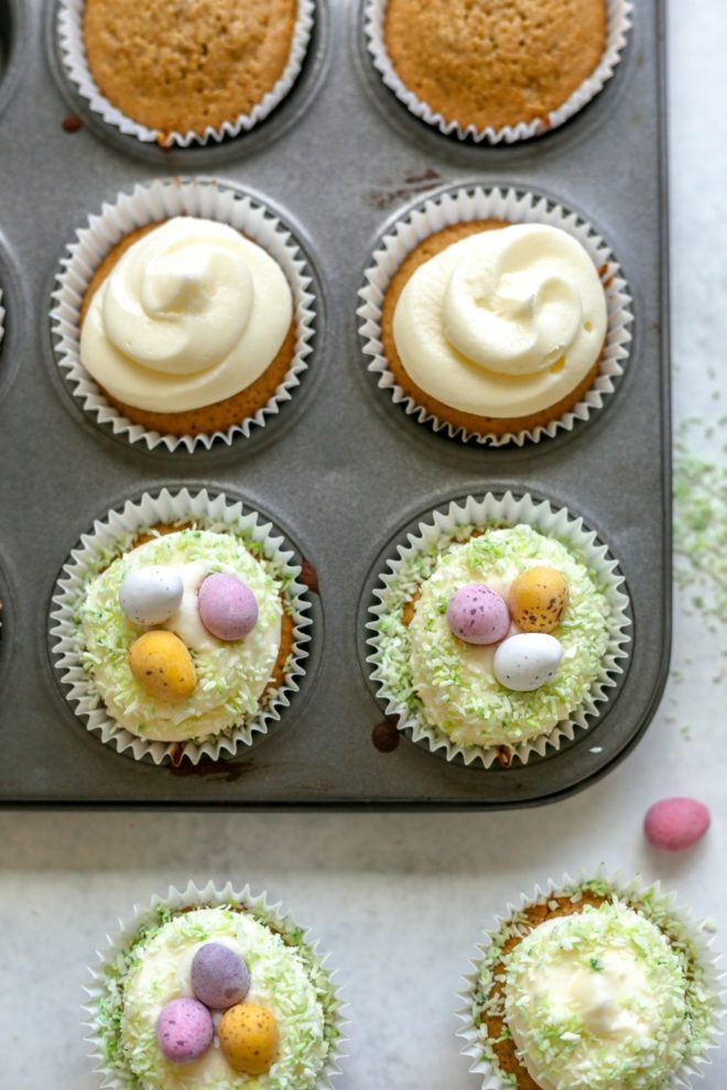 This is an overhead image of vanilla cupcakes. Some cupcakes are in the cupcake tin and some are on the white surface at the bottom of the image. The cupcakes are topped with vanilla frosting, sprinkled with green coconut and mini candy eggs.
