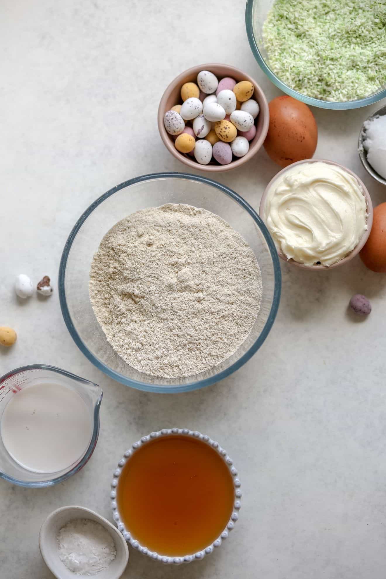 This is an overhead image of ingredients to make vanilla cupcakes with mini easter candy eggs on top. The ingredients are on a white surface.