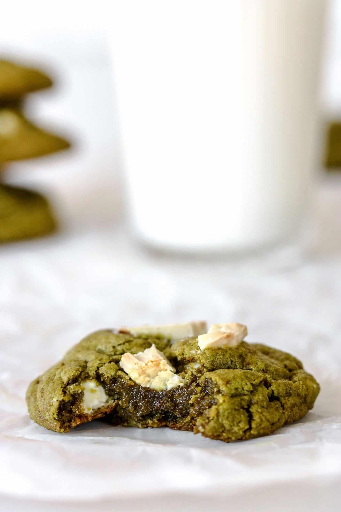 This is a side view of a green cookie with white chocolate chunks and a bite taken out of it. The cookies sit on a white piece of parchment paper with a glass of milk and more green cookies blurred in the background.