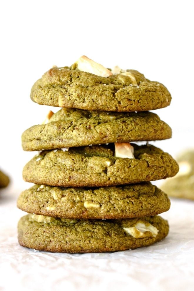 This is a stack of five green matcha cookies sitting on a white piece of parchment paper. More cookies are blurred in the background.