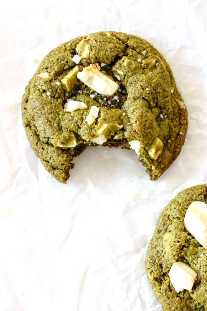 This is an overhead image of a green matcha cookie with white chocolate chunks. A bite is taken out of the cookie and is sitting on a white piece of parchment paper. Another cookie is in the bottom right corner of the image.