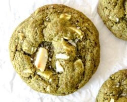 This is an overhead image of green cookies with white chocolate cunks on top. The cookies are sprinkled with some flakey salt and sitting on a white piece or parchment paper. Text overlay reads "white chocolate matcha cookies."