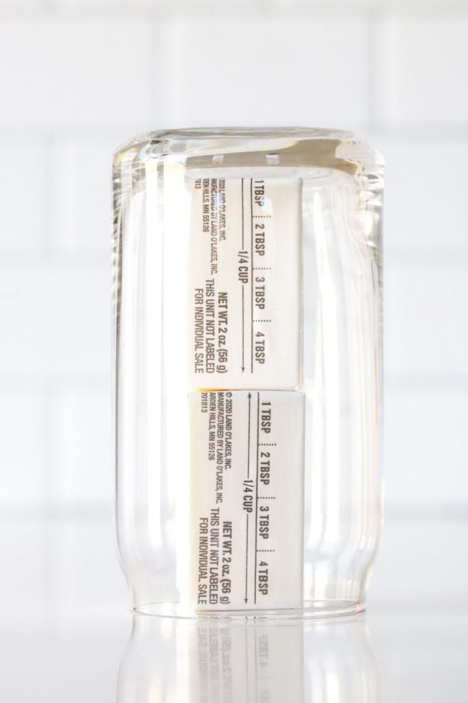 This is a side view of a glass upside down over a stick of butter. The glass and butter sit on a white counter with a white tile background.