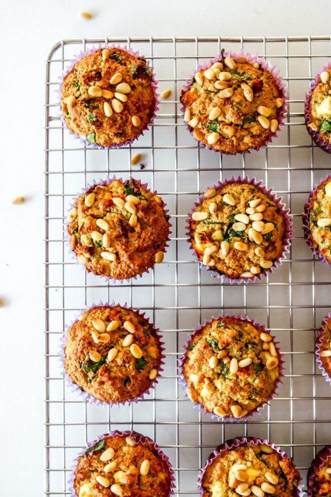 This is an overhead image of a cooling rack with spinach and feta muffins. The muffins are topped with pine nuts. The cooling rack sits on a white counter.