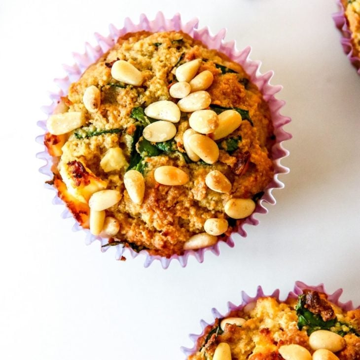 This is an overhead image of muffins with pine nuts and spinach. The muffin is lined with a purple paper liner and sits on a white counter with two other muffins to the right of the images and a couple pine nuts on the counter.