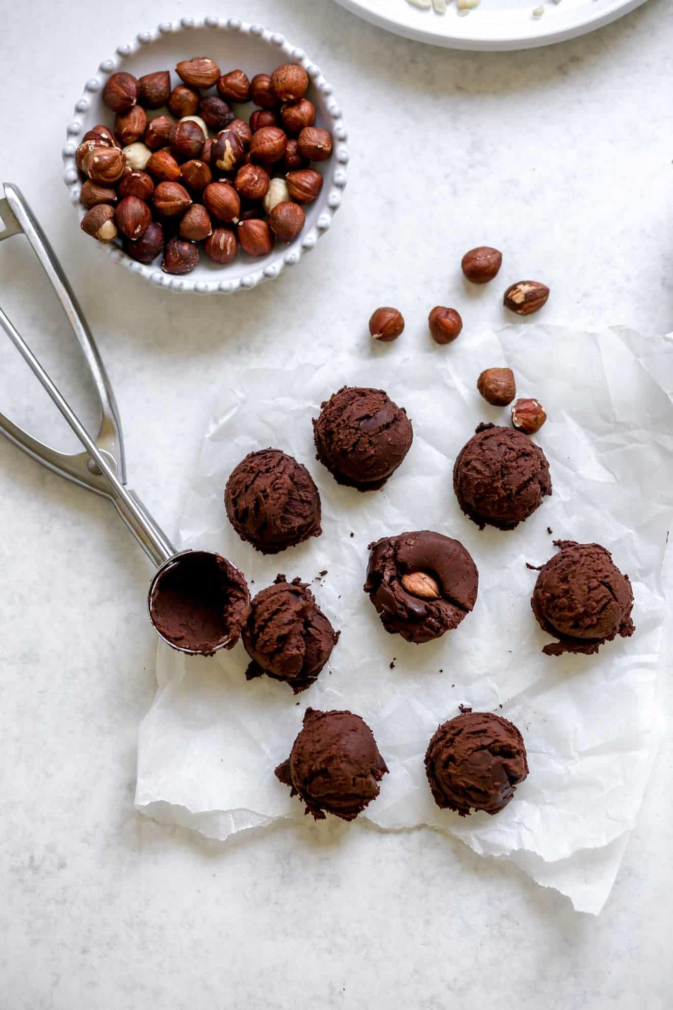 This is an overhead image looking to chocolate truffles on a white piece of parchment paper on a light grey surface. The chocolate truffles have a cookie scooper laying next to them. The center chocolate ball has a hazelnut in the middle. To the top left is a small bowl of hazelnuts.
