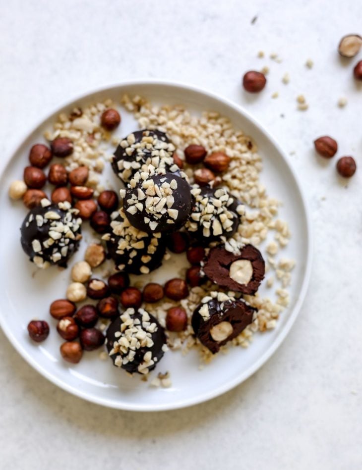 This is an overhead view of a white plate with chocolate truffles coated in chopped nuts. One candy is cut in half and inside is a hazelnut. The white plate is on a light grey surface with some whole hazelnuts off to the top left of the image.