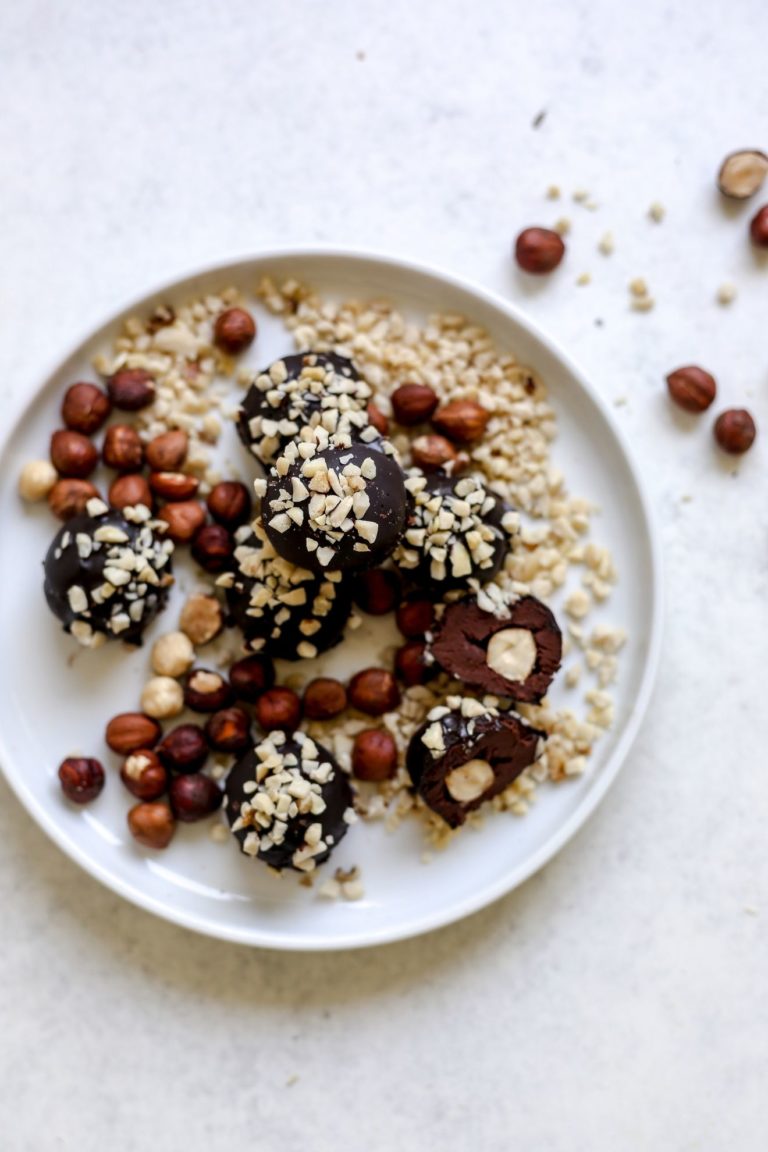 This is an overhead view of a white plate with chocolate truffles coated in chopped nuts. One candy is cut in half and inside is a hazelnut. The white plate is on a light grey surface with some whole hazelnuts off to the top left of the image.