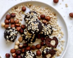 This is an overhead view of a white plate with chocolate truffles coated in chopped nuts. One candy is cut in half and inside is a hazelnut. The white plate is on a light grey surface with some whole hazelnuts off to the top left of the image. Text overlay reads "vegan ferrero rocher."