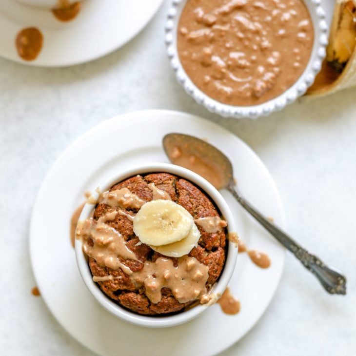 This is an overhead image of a ramekin with baked oats. The blended baked oats are topped with a drizzle of peanut butter and sliced banana. The ramekin is on a white plate with an antique spoon with peanut butter on it. The plate sits on a light grey surface. Another ramekin with baked oats is in the top left corner of the image. A small white bowl of peanut butter and an open, sliced banana is in the top right corner of the image.