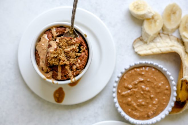 This is an overhead image of a ramekin with baked oats. The blended baked oats are topped with a drizzle of peanut butter and the ramekin is on a white plate. An antique spoon is in the baked oats. The plate sits on a light grey surface. A small white bowl of peanut butter and an open, sliced banana is to the right of the image.