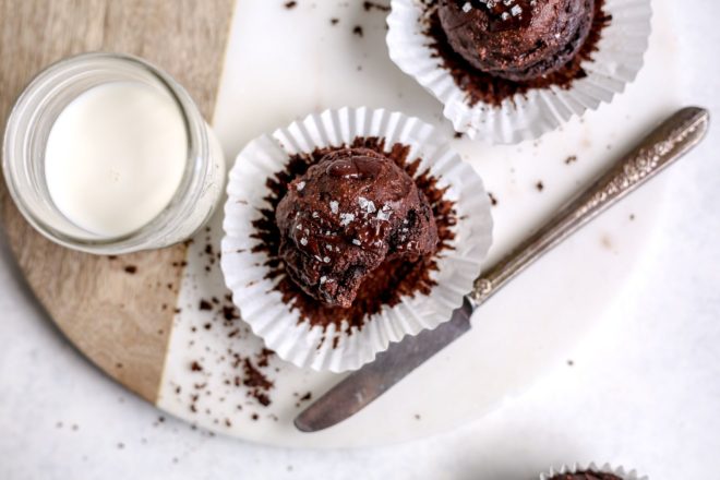 This is an overhead image of a chocolate muffin with the muffin liner pulled away. The muffin is sprinkled with salt and sits on a wood and marble cutting board. Another muffin is to the top right of the main muffin and a glass of milk to the left.