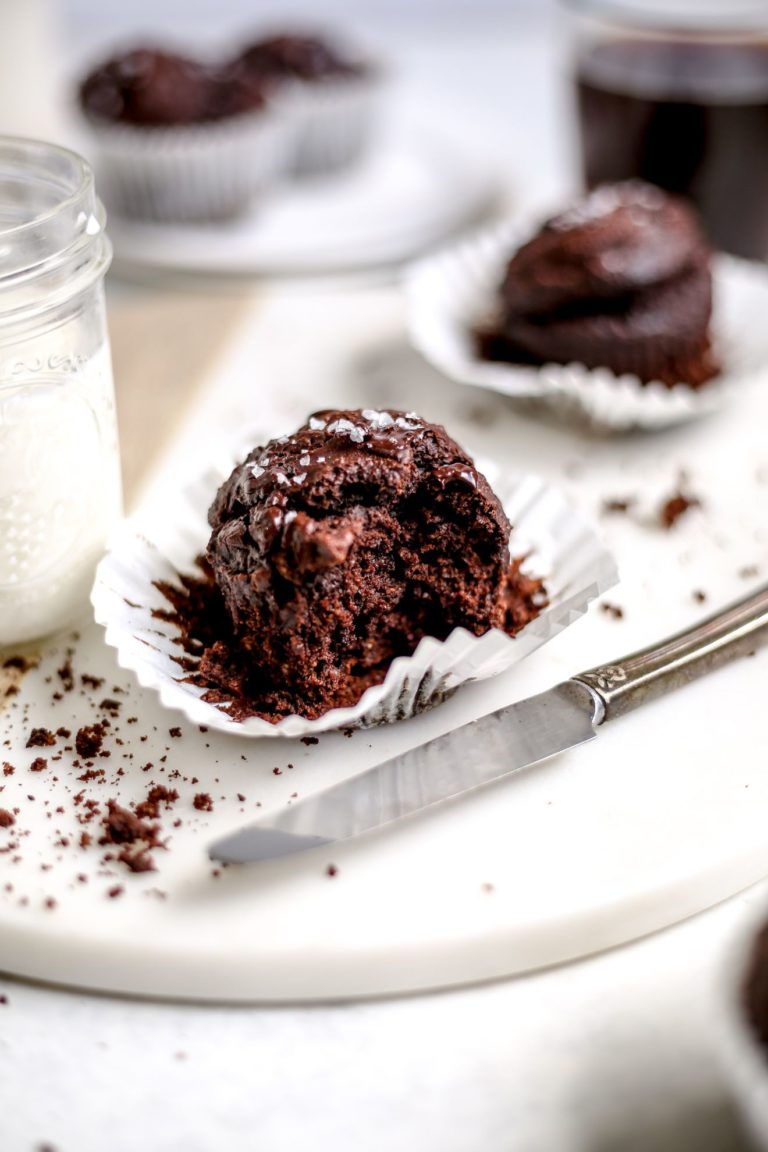 This is a side view of a chocolate muffin with the muffin liner pulled away. The muffin sits on a cutting board and a bite is taken out of it. More muffins blurred in the background. A glass of milk is to the left of the muffin.