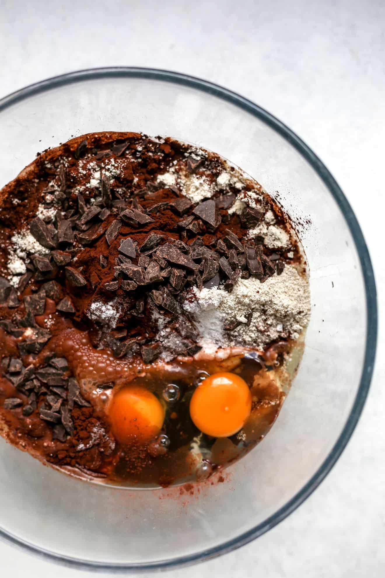 This is an overhead image of a glass bowl with eggs, chocolate chips, cocoa, oat flour, and other ingredients. The glass bowl sits on a white surface.
