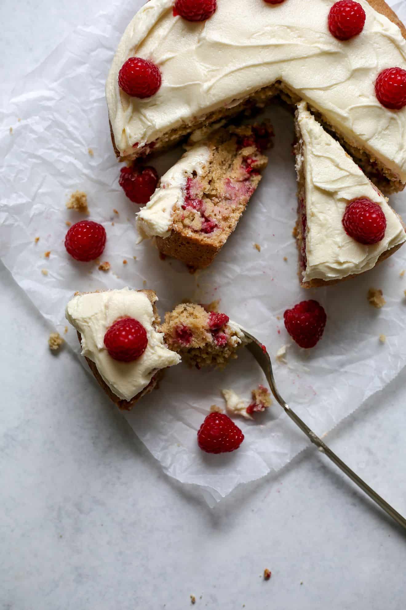 This is an overhead image of a vanilla raspberry cake topped with vanilla icing and fresh raspberries. The cake sits on a white piece of parchment paper on a white counter. Slice of the cake are cut and one has a fork taking bites from the slice.