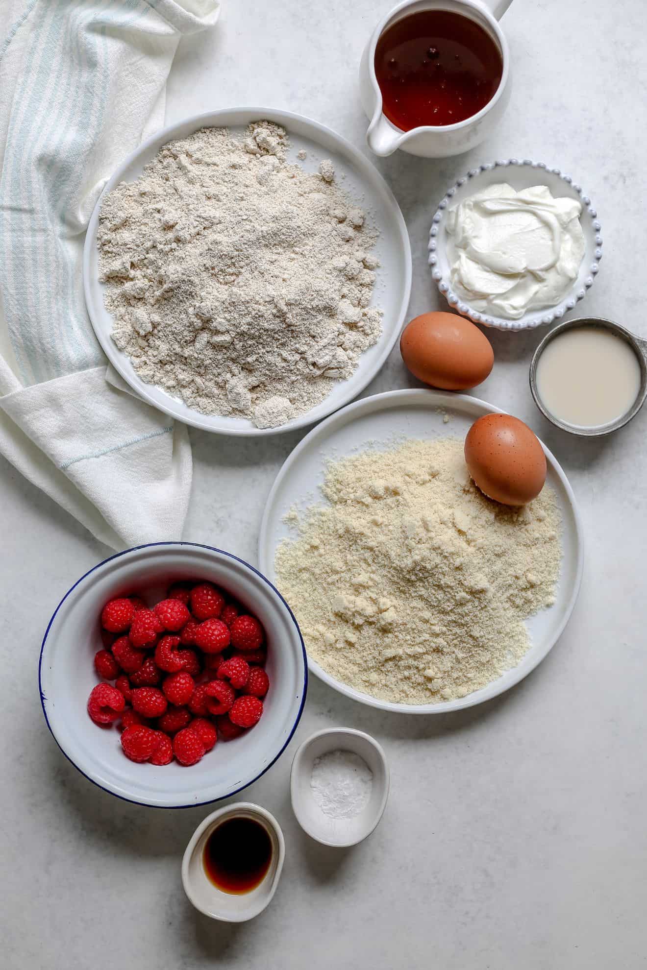 This is an overhead image of the ingredients needed for a raspberry cake. The oat flour, almond flour, yogurt, raspberries, eggs and other ingredients are on a white background.