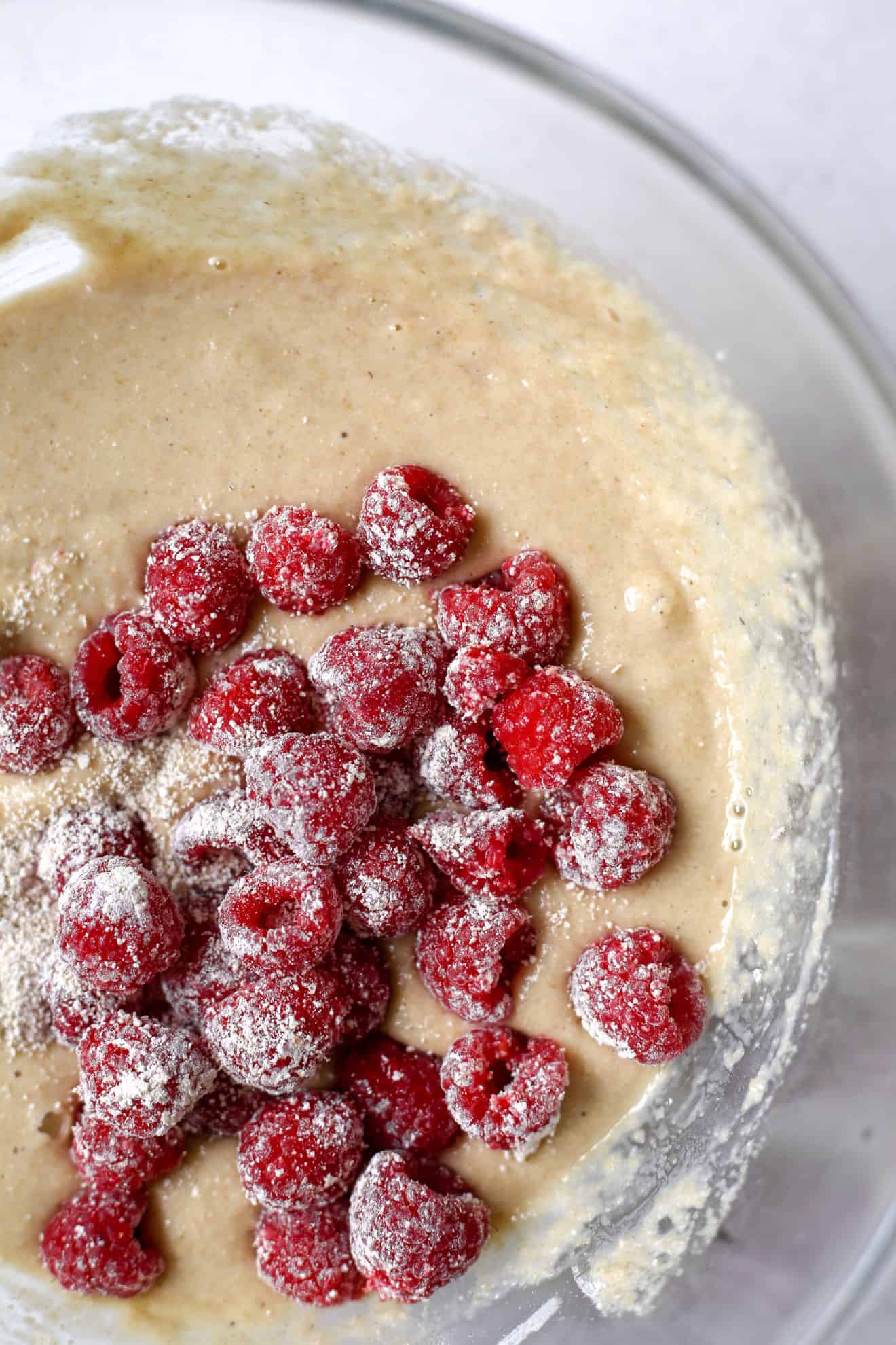 This is an overhead image of a glass bowl with raw batter inside. Fresh raspberries coated with flour are on top of the raw batter. The glass bowl sits on a white counter.