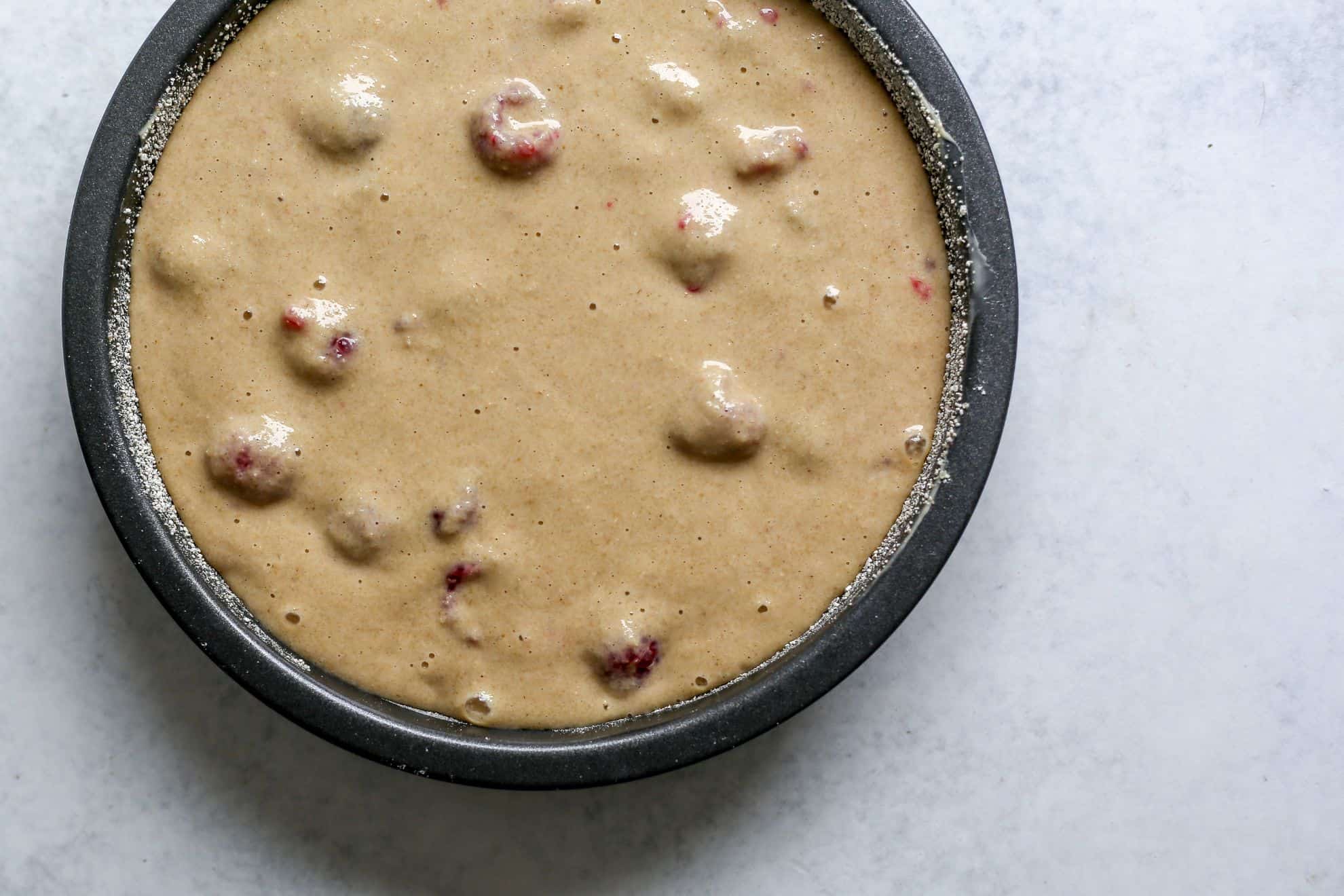 This is an overhead image of a circle pan with raw raspberry cake batter in it. You can see the raw raspberries coated with batter. The pan sits on a white counter.