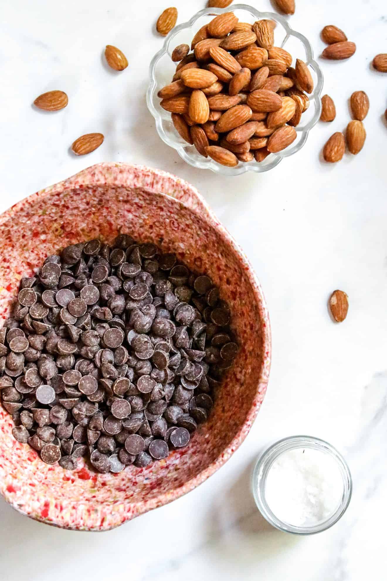 This is an overhead image of a small glass bowl with almonds, a larger pink speckled bowl with chocolate chips and a small glass jar with flakey salt. All the items sit on a marble countertop.