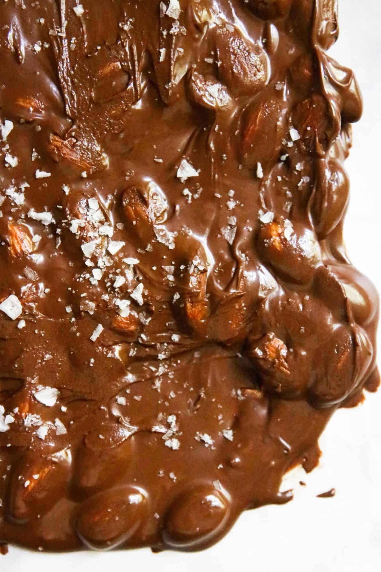 This is an overhead image of melted chocolate with almonds spread out on a white piece of parchment paper. The chocolate is sprinkled with flakey salt.