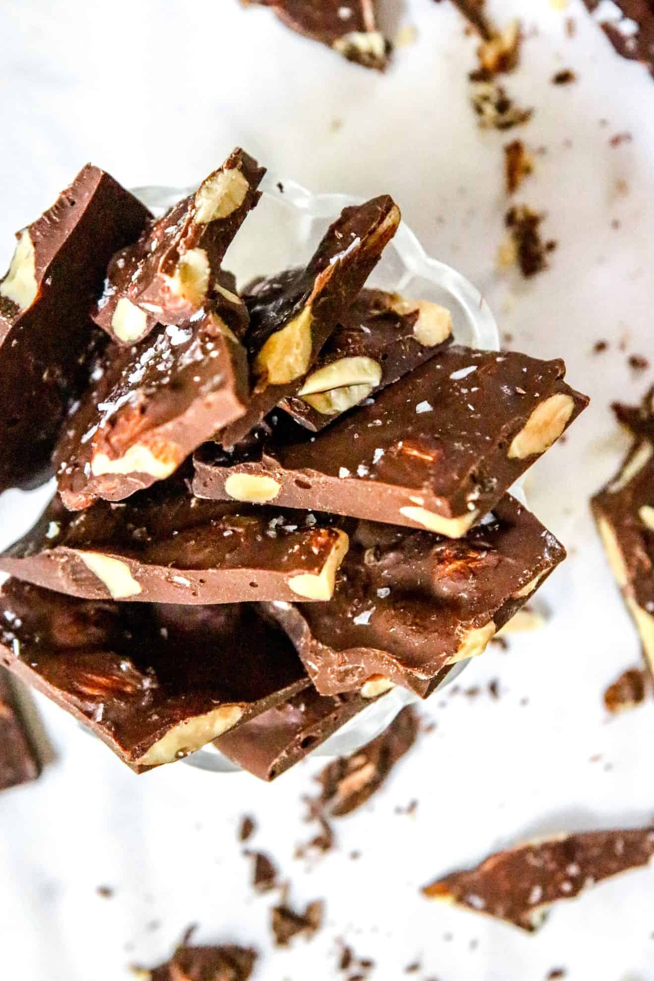 This is an overhead image of chocolate bark with almonds and flakey salt on top. The bark pieces are in a small glass bowl sitting on a white marble counter. Small pieces of bark and crumbs are scattered around the bowl.