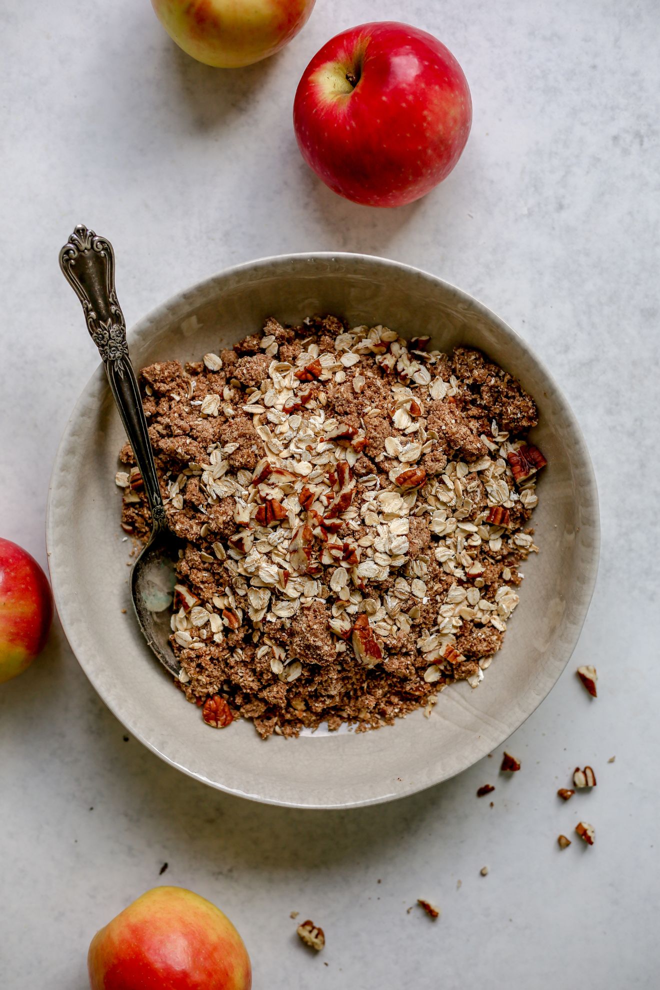 This is an overhead image of a beige bowl with an oat pecan crumble mixture in it. An antique spoon is in the bowl leaning against the side. The bowl sits on a white gray surface with apples scattered around.