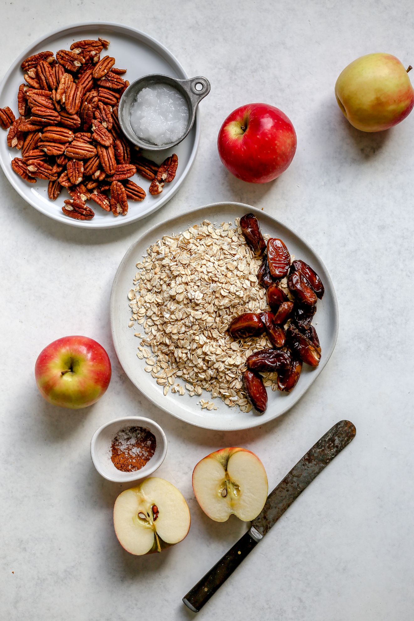 This is an overhead image of plates and bowls with assorted ingredients: pecans, coconut oil, apples, oats, dates, and spices. There is an apple cut in half toward the bottom of the image with an antique butter knife