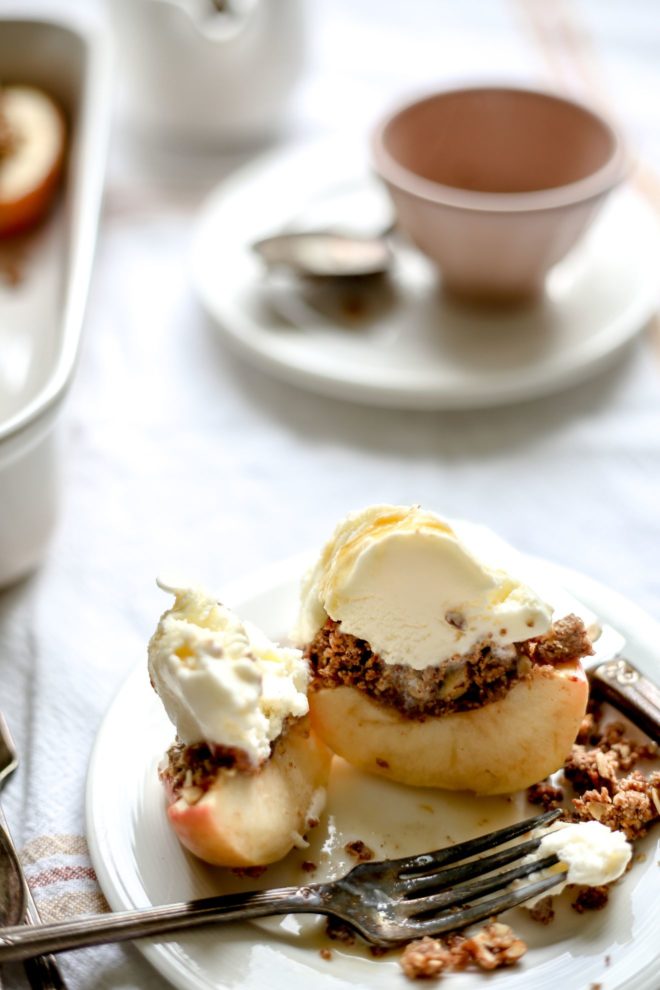 This is a side view of a baked apple cut in half. The apple is topped with a cinnamon oat crumble and vanilla ice cream. The apple is cut into and sits on a white plate with a fork. The plate sits on a white table cloth and a baking dish with more apples are to the left of the image. A teacup are blurred in the background.