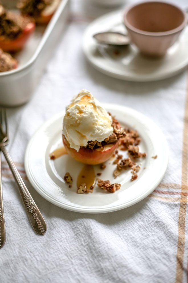 This is an overhead angled image of a baked apple on a white plate. The baked apple is topped with a crumble, vanilla ice cream and caramel drizzle. The plate is on a white table cloth and a baking dish with more baked apples are to the upper left of the image, blurred in the background along with a pink teacup to the right.