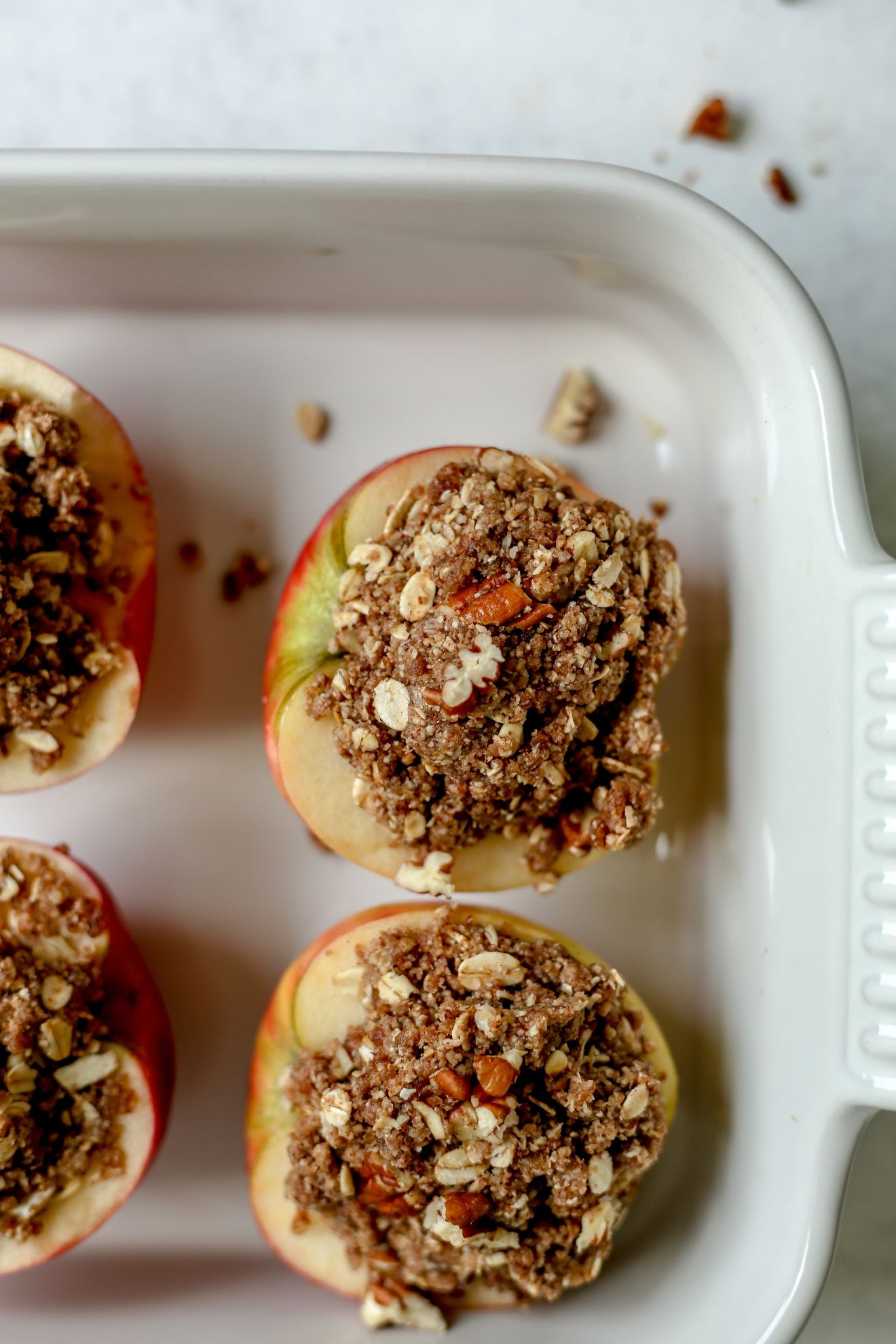 This is an overhead image of a baking dish with halved apples and topped with an oat crumble. The backing dish has four apples in it and is sitting on a white surface.