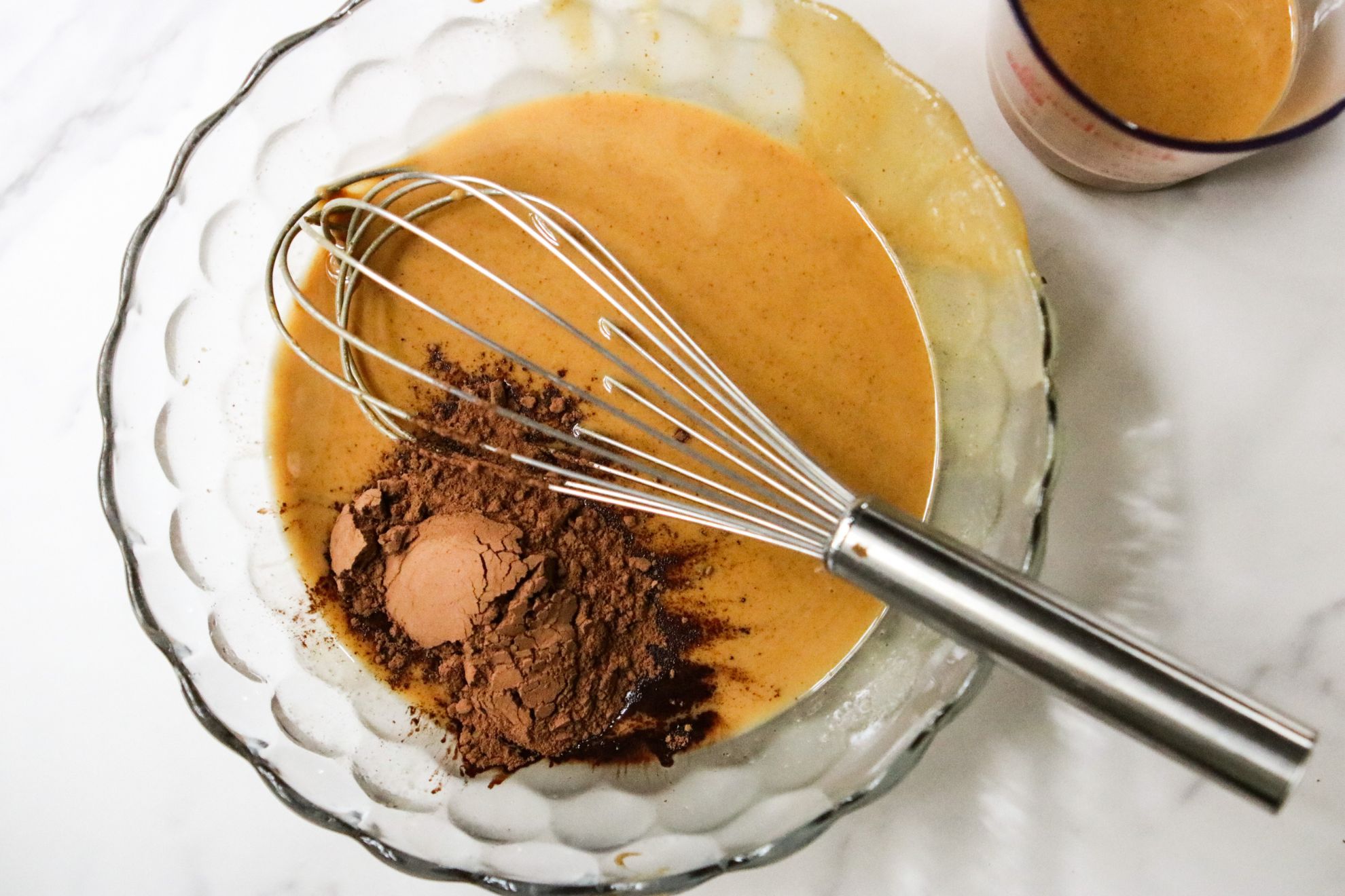 This is an overhead image of a glass bowl on a white counter. Inside the bowl is melted peanut butter and cocoa powder and a whisk. Off to the top right of the image is a measuring cup with some of the peanut butter mixture.