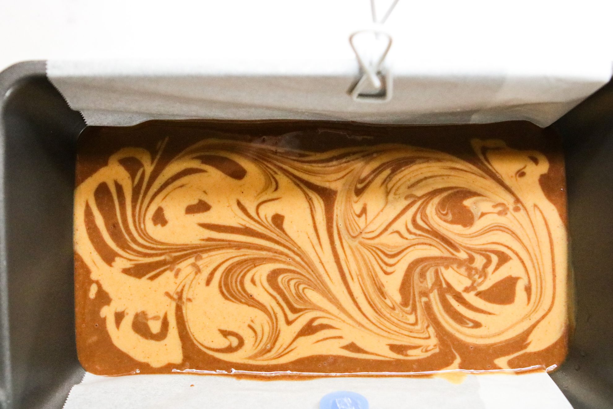 This is an overhead image of a bread pan with parchment paper sitting on a white counter. Inside the bread pan is a chocolate mixture with peanut butter swirls.
