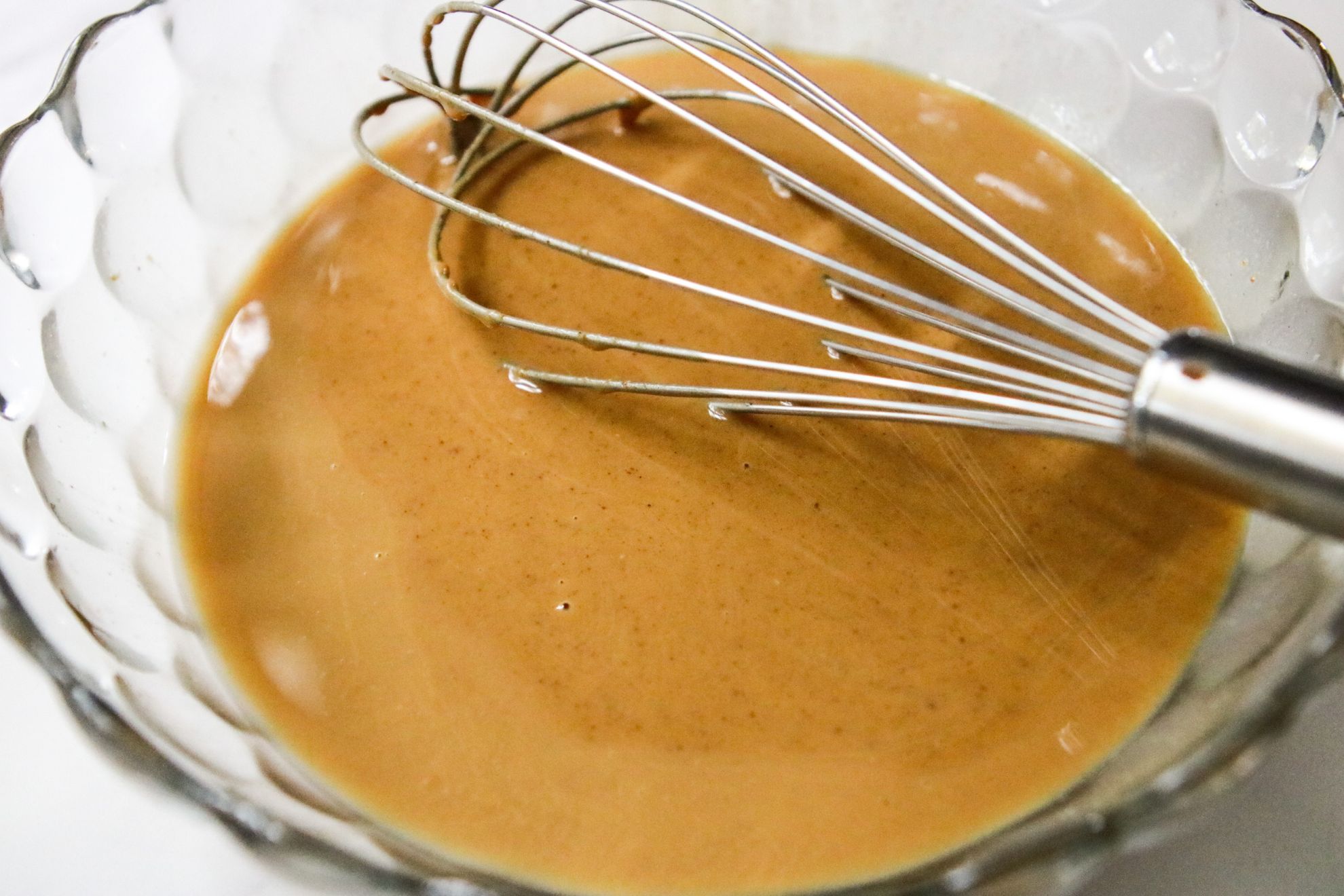 This is a side view of a glass bowl sitting on a white counter. In the bowl is a peanut butter mixture with a whisk.