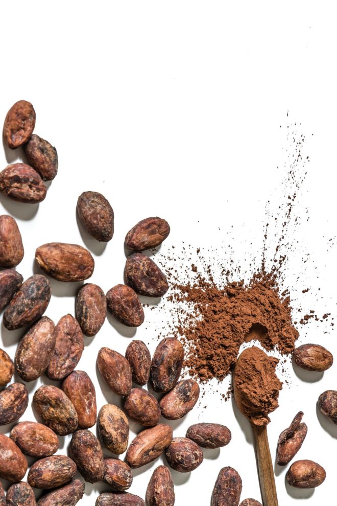 This is an overhead image of cacao beans on a white surface. A spoon has cocoa powder on it and is splattering onto the white surface.