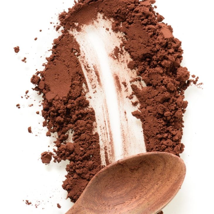 This is an image of cocoa powder being smeared on a white surface. A wood spoon rests in the powder, leaving a trail of powder brushed away behind it.