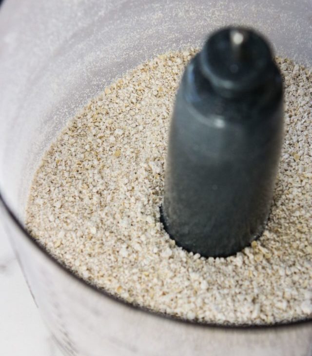 This is an overhead angled shot looking down into a food processor with oat flour in it. The food processor sits on a white marble countertop.