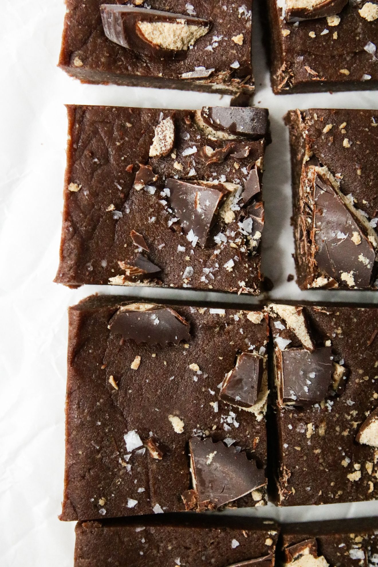 This is an overhead image of brownies cut into squares. The brownies are topped with cut up peanut butter cups and flakey salt. The brownies are on a white piece of parchment paper.