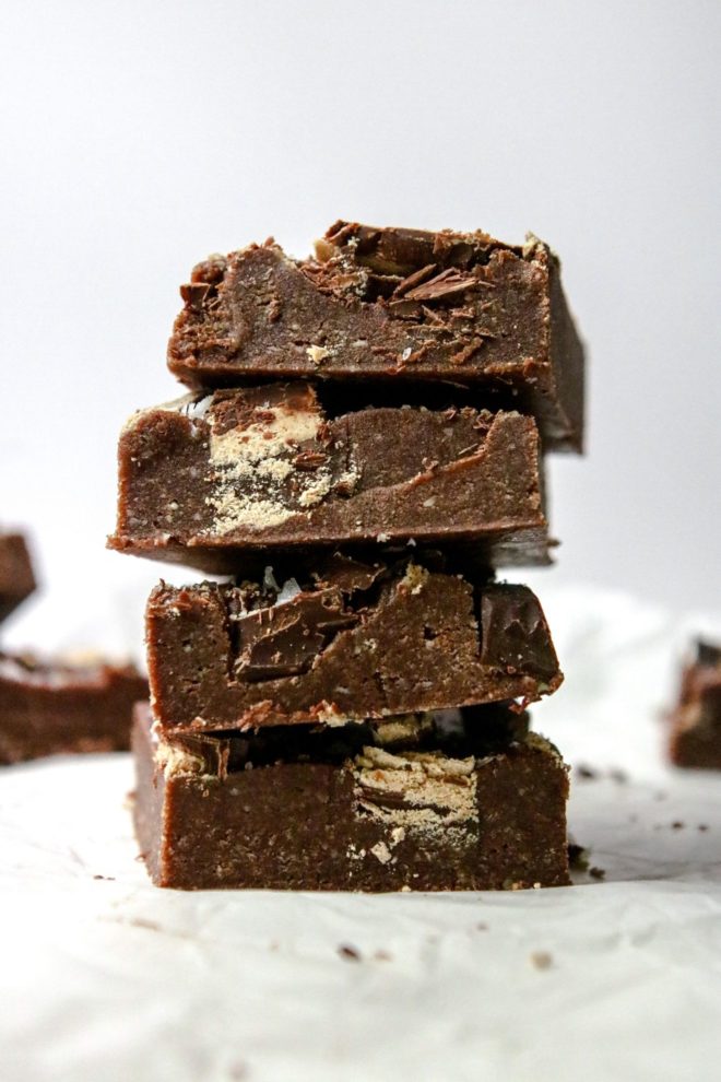 This is a side view of a stack of four no bake chocolate brownies sitting on a white piece of parchment paper. More brownies are blurred in the background.