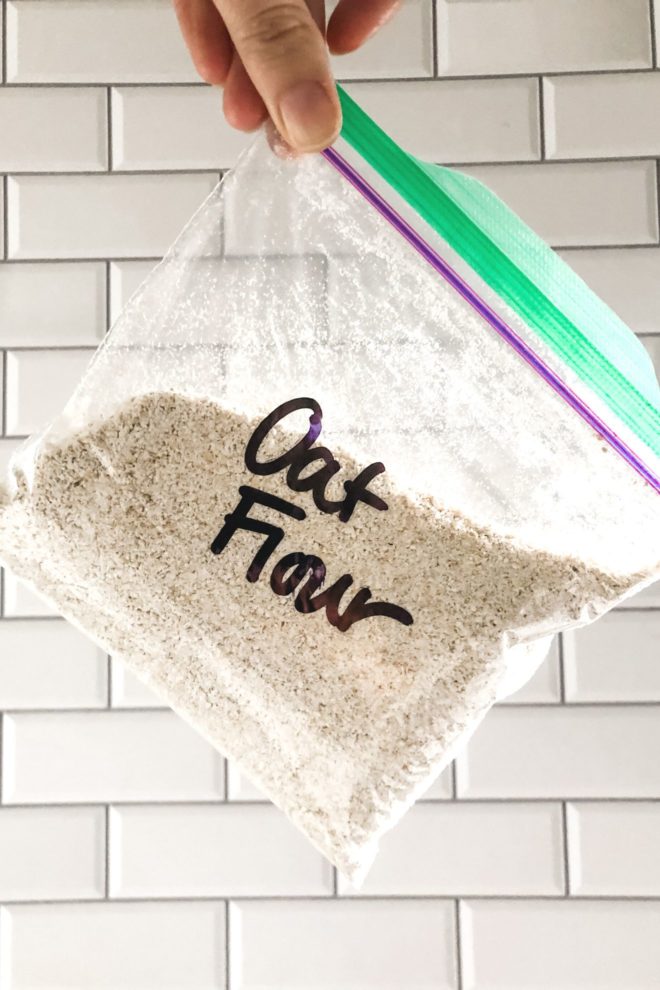 This is a vertical image of a hand holding a baggie filled with a beige powder. "Oat Flour" is written in black sharpie on the plastic bag. White subway tile is in the background.