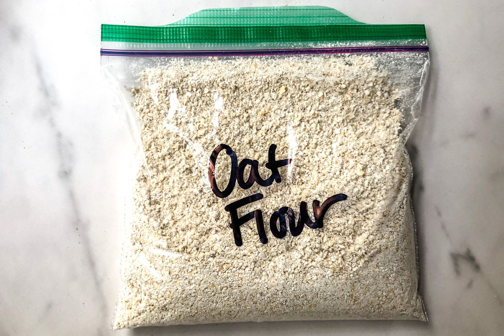 This is an overhead image looking onto a plastic baggie filled with oat flour. "Oat Flour" is written on the baggie in black sharpie. The bag lays on a white marble surface.