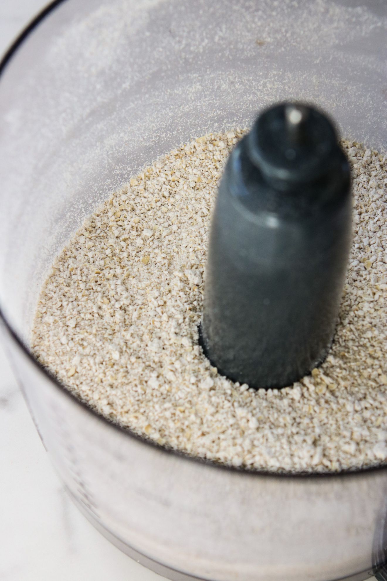 This is an overhead angled shot looking down into a food processor with oat flour in it. The food processor sits on a white marble countertop.