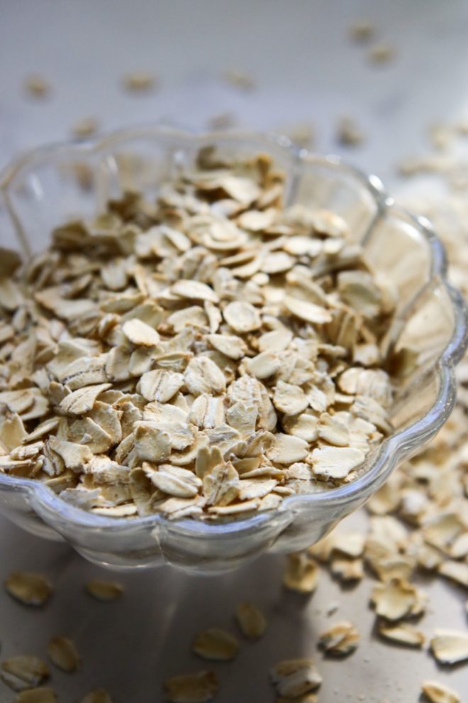 This is a side view of a small scalloped glass bowl filled with oats. More oats are scattered around the bowl on the white counter.