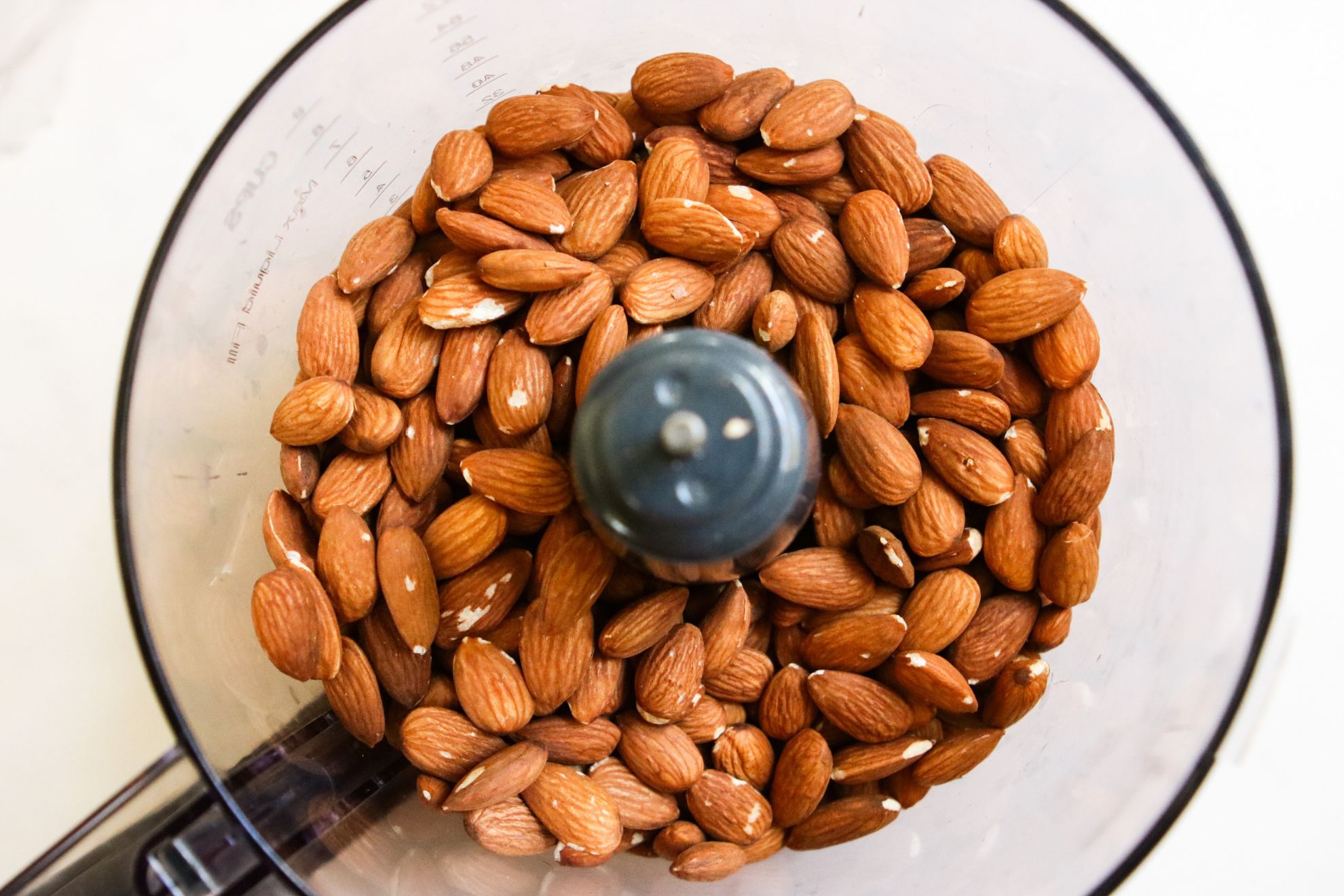 This is an overhead horizontal image of almonds in a food processor. The food processor sits on a white marble surface.