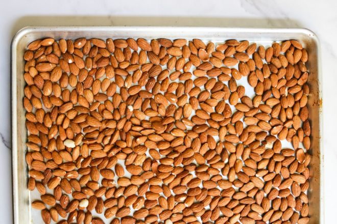 This is an overhead image of almonds spread out on a rimmed baking sheet. The baking sheet sits on a white marble counter.